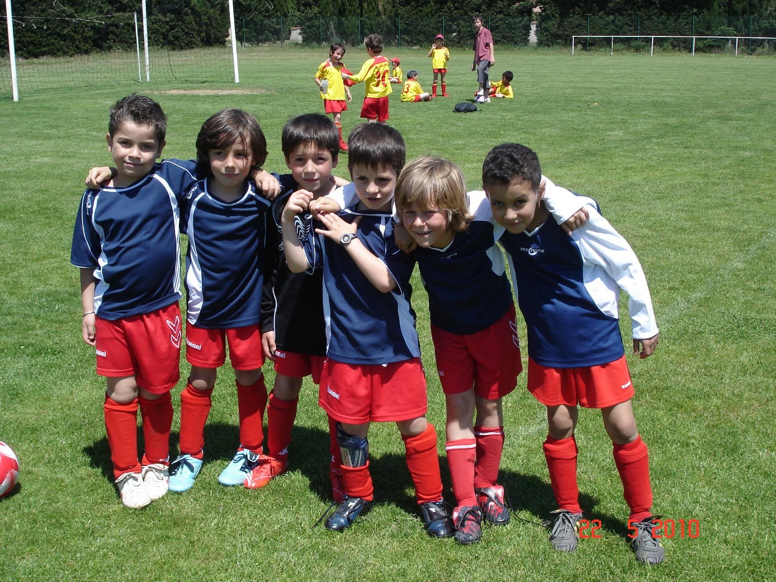 A children's football team lined up arm in arm on the pitch, in navy and red kit, after a match.