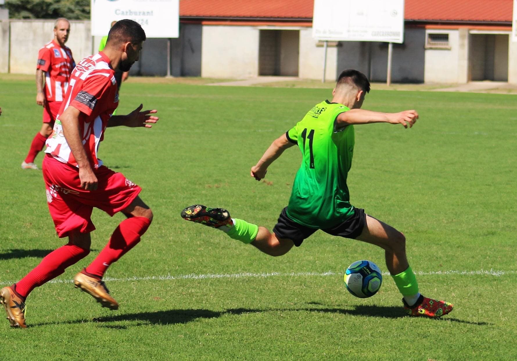 A player in a green kit striking the ball at full stretch during a match, chased by a defender in red.