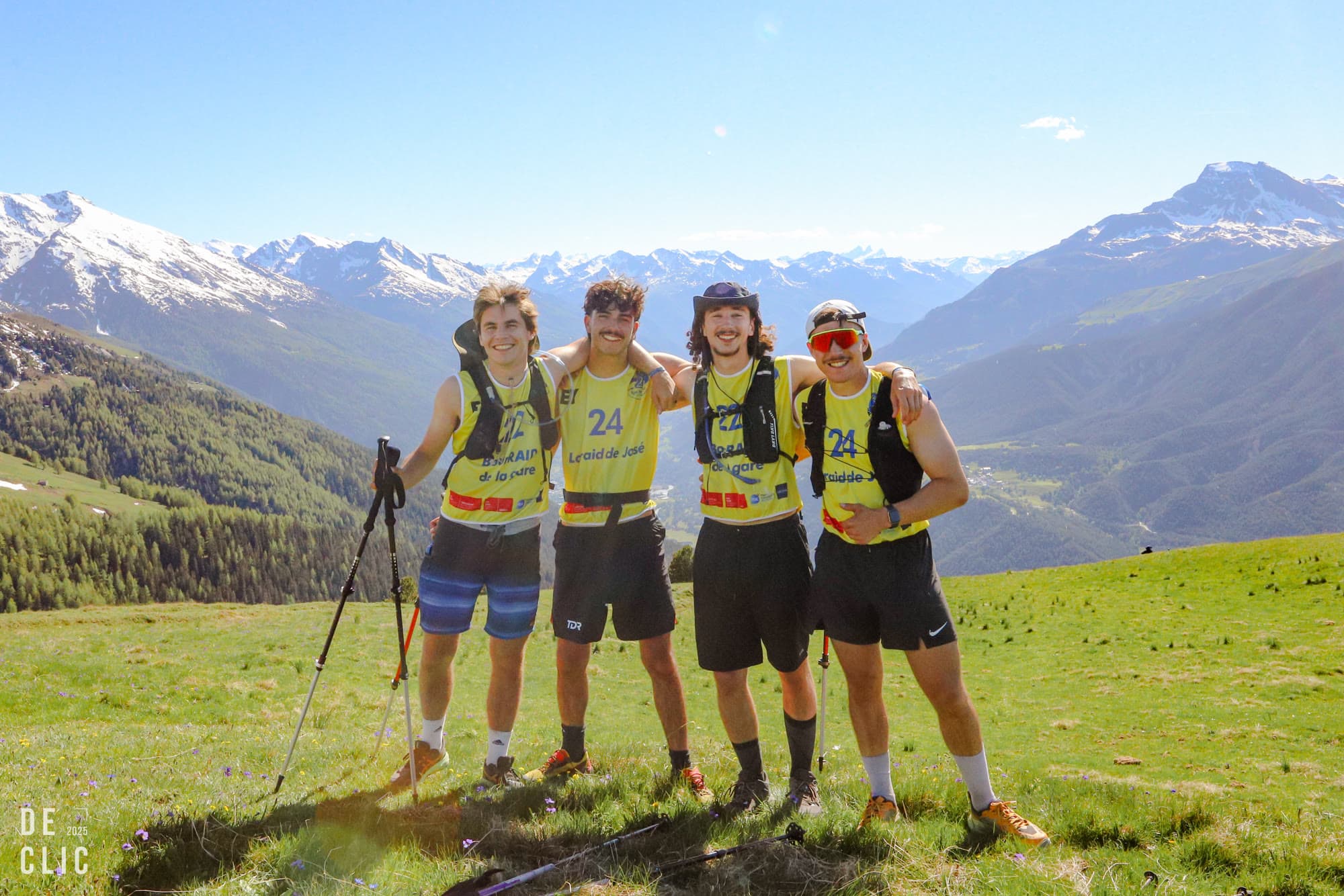 Four runners in yellow race shirts standing arm in arm on an alpine meadow, snow peaks behind.