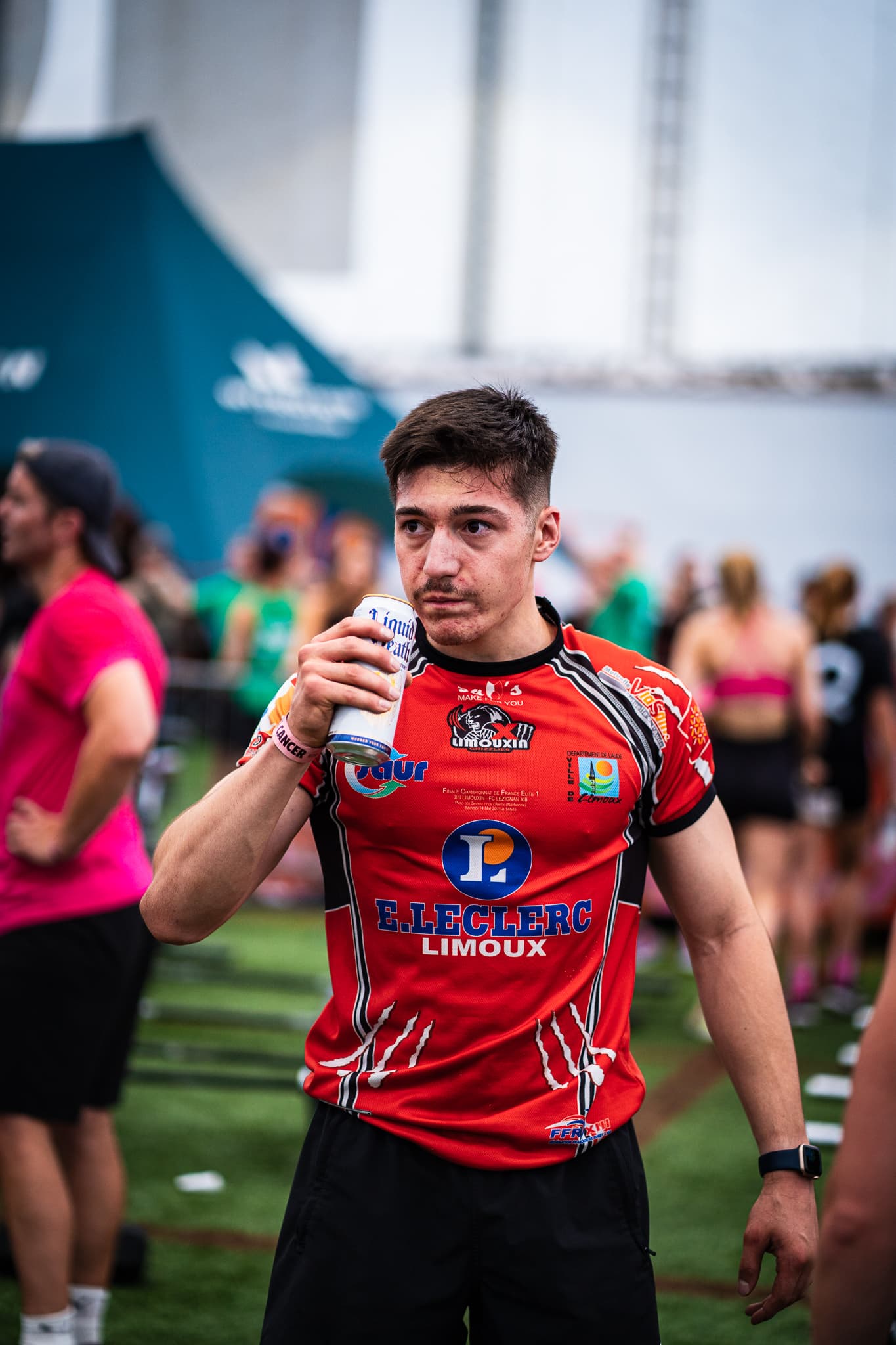 Standing on a grass field in a red E.Leclerc Limoux shirt, a can raised, other athletes behind.
