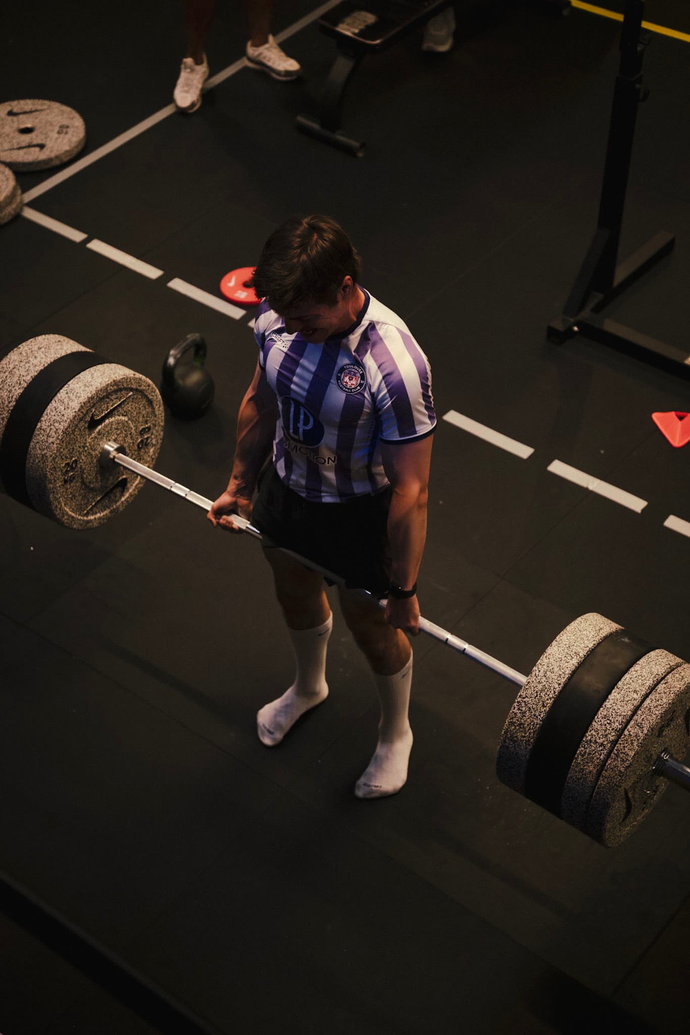 Lifting a loaded barbell in a gym while wearing a football shirt and match socks.